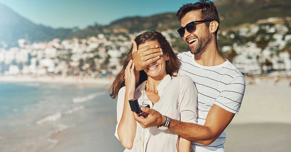 Mann verdeckt Frau Augen am Strand und hat Verlobungsring in der Hand