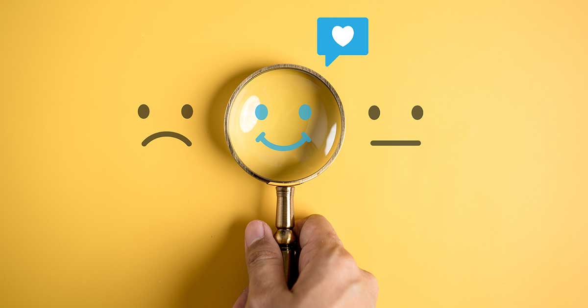 Hand hält eine Lupe über ein lachendes Gesicht zwischen einem traurigen und einem neutralen Smiley – Fokus auf positive Stimmung.