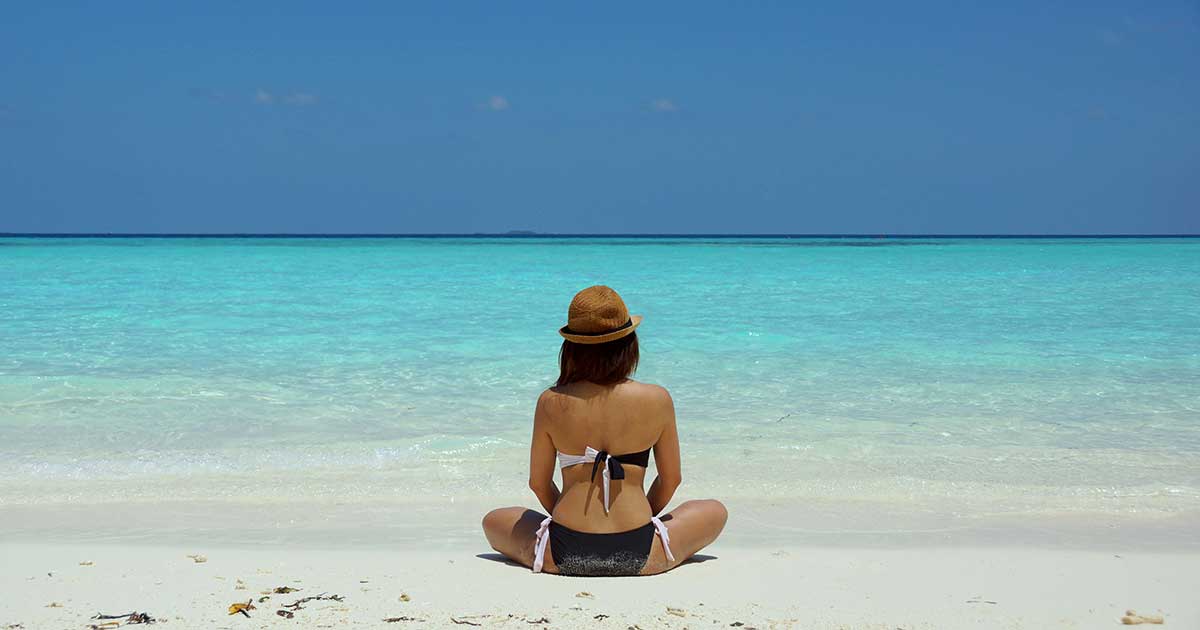 Frau im Bikini mit Strohhut sitzt am Strand und blickt aufs türkisblaue Meer