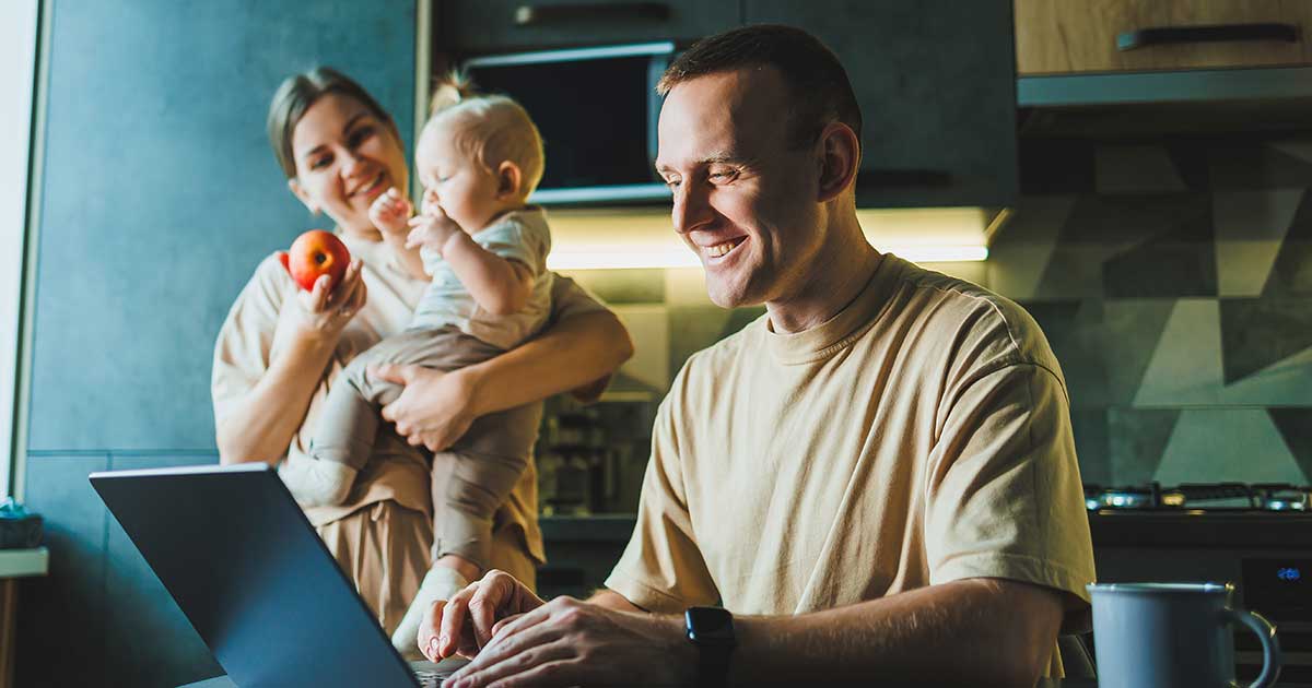 Vater arbeitet am Laptop in der Küche, während Mutter mit Baby im Hintergrund beschäftigt ist – typische Alltagssituation beim Wiedereinstieg in den Beruf.