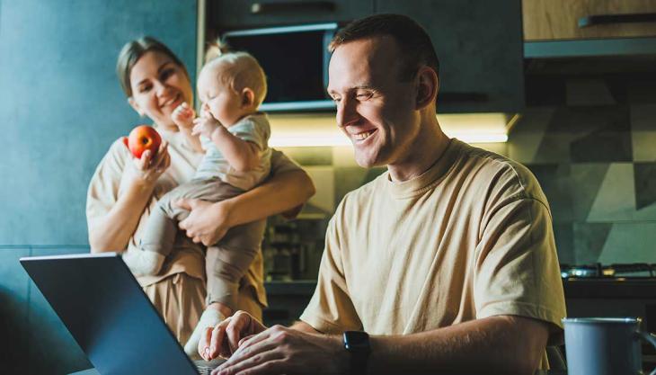 Vater arbeitet am Laptop in der Küche, während Mutter mit Baby im Hintergrund beschäftigt ist – typische Alltagssituation beim Wiedereinstieg in den Beruf.