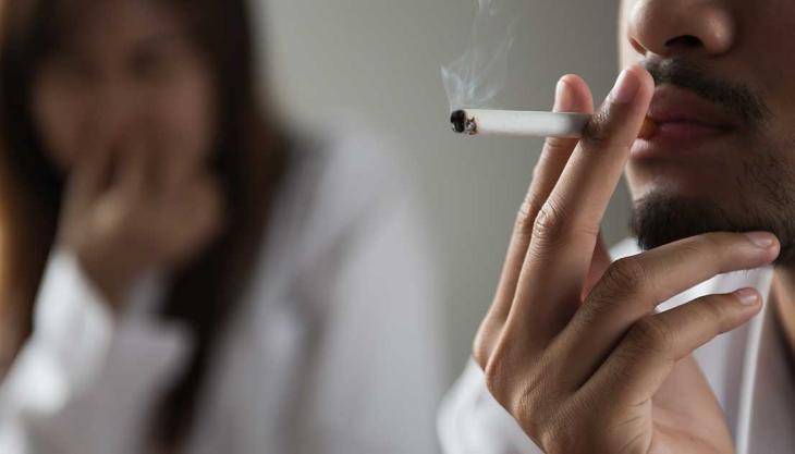 Mann raucht eine Zigarette, Partnerin im Hintergrund wirkt genervt und distanziert