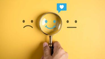 Hand hält eine Lupe über ein lachendes Gesicht zwischen einem traurigen und einem neutralen Smiley – Fokus auf positive Stimmung.