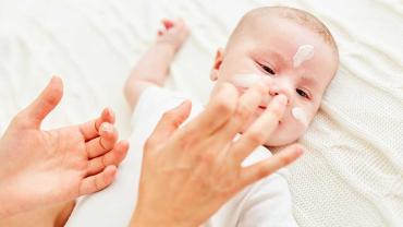 Mutter trägt einem Baby sanft Creme auf die empfindliche Gesichtshaut auf
