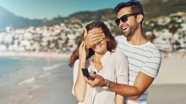 Mann verdeckt Frau Augen am Strand und hat Verlobungsring in der Hand