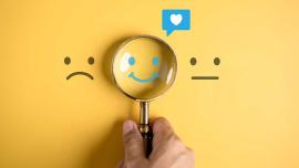 Hand hält eine Lupe über ein lachendes Gesicht zwischen einem traurigen und einem neutralen Smiley – Fokus auf positive Stimmung.