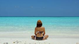 Frau im Bikini mit Strohhut sitzt am Strand und blickt aufs türkisblaue Meer