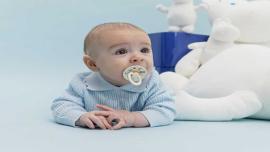 Baby mit Schnuller im Mund liegt auf einer hellblauen Unterlage neben weißen Kuscheltieren.