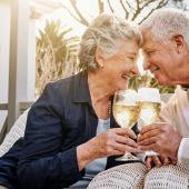 Älteres Ehepaar stößt mit Sektgläsern an und feiert gemeinsam auf der Terrasse ihre Goldene Hochzeit – beide lächeln sich liebevoll an.