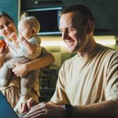 Vater arbeitet am Laptop in der Küche, während Mutter mit Baby im Hintergrund beschäftigt ist – typische Alltagssituation beim Wiedereinstieg in den Beruf.
